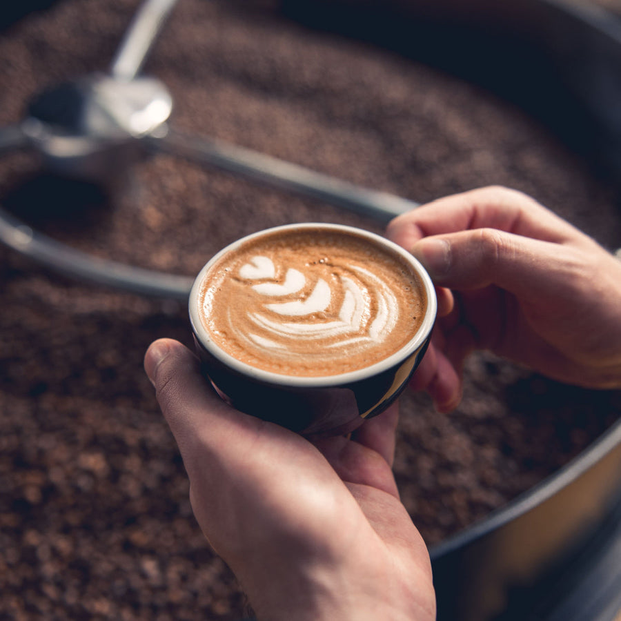 A person holds a small cup of latte art with a leaf design, surrounded by a background of coffee beans and equipment, suggesting a coffee preparation setting.