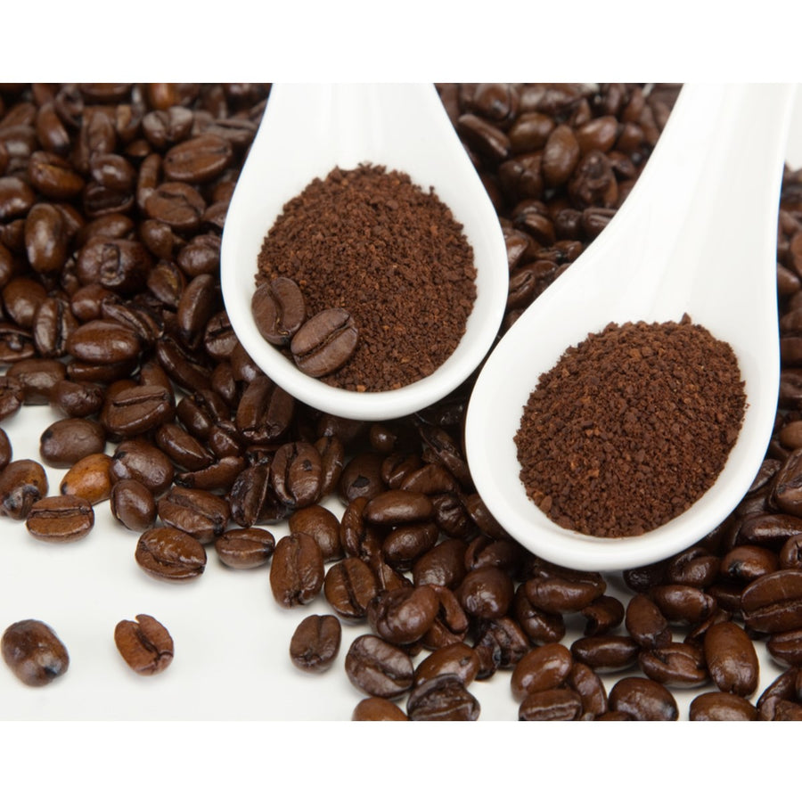 Two white spoons hold ground coffee and whole coffee beans, surrounded by scattered roasted coffee beans on a white surface, showcasing the different forms of coffee.