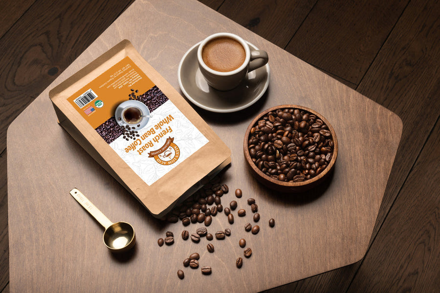 A bag of freshly roasted whole bean coffee sits on a wooden table next to a small cup of coffee, a bowl of coffee beans, and a gold measuring spoon, with some beans scattered around.
