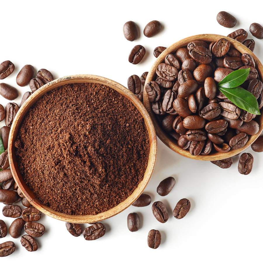 Two wooden bowls contain ground coffee and whole coffee beans, with scattered beans and a green leaf nearby, set against a white background.