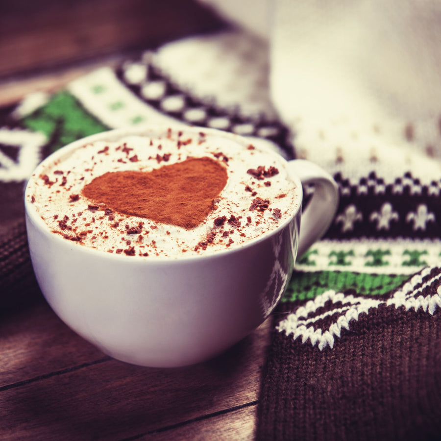 A white cup of hot chocolate topped with whipped cream and cocoa powder shaped like a heart sits on a cozy, patterned knitted blanket.