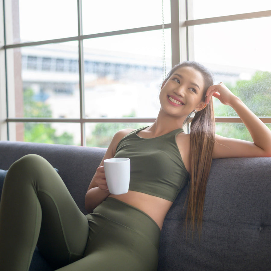 A woman in olive green activewear sits on a gray sofa, smiling and holding a white mug, with large windows and greenery visible in the background.