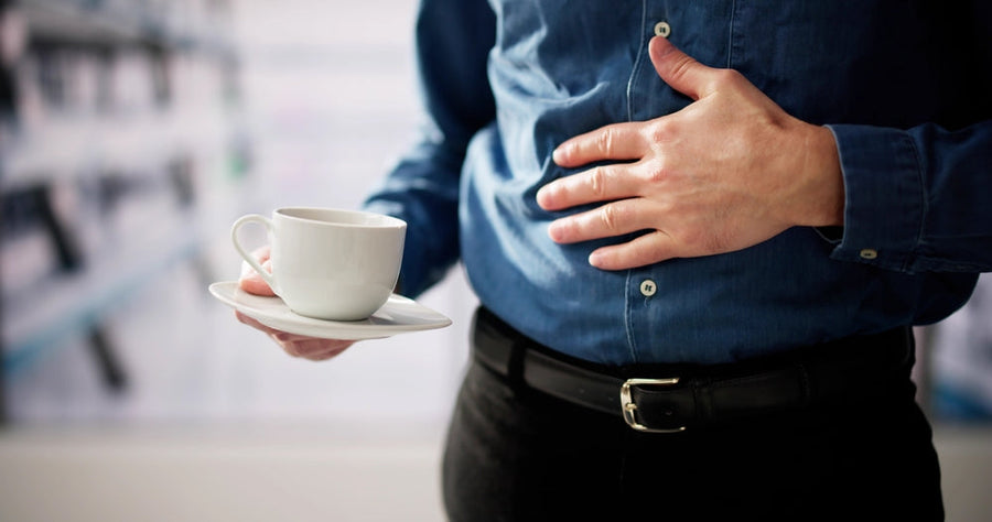 A person in a blue shirt holds a white cup on a saucer in one hand while resting their other hand on their stomach, appearing contemplative in a blurred indoor setting.
