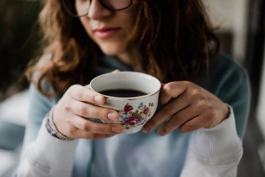 A young woman with curly hair and glasses gently holds a floral-patterned cup of coffee, appearing contemplative while seated indoors in a cozy setting.