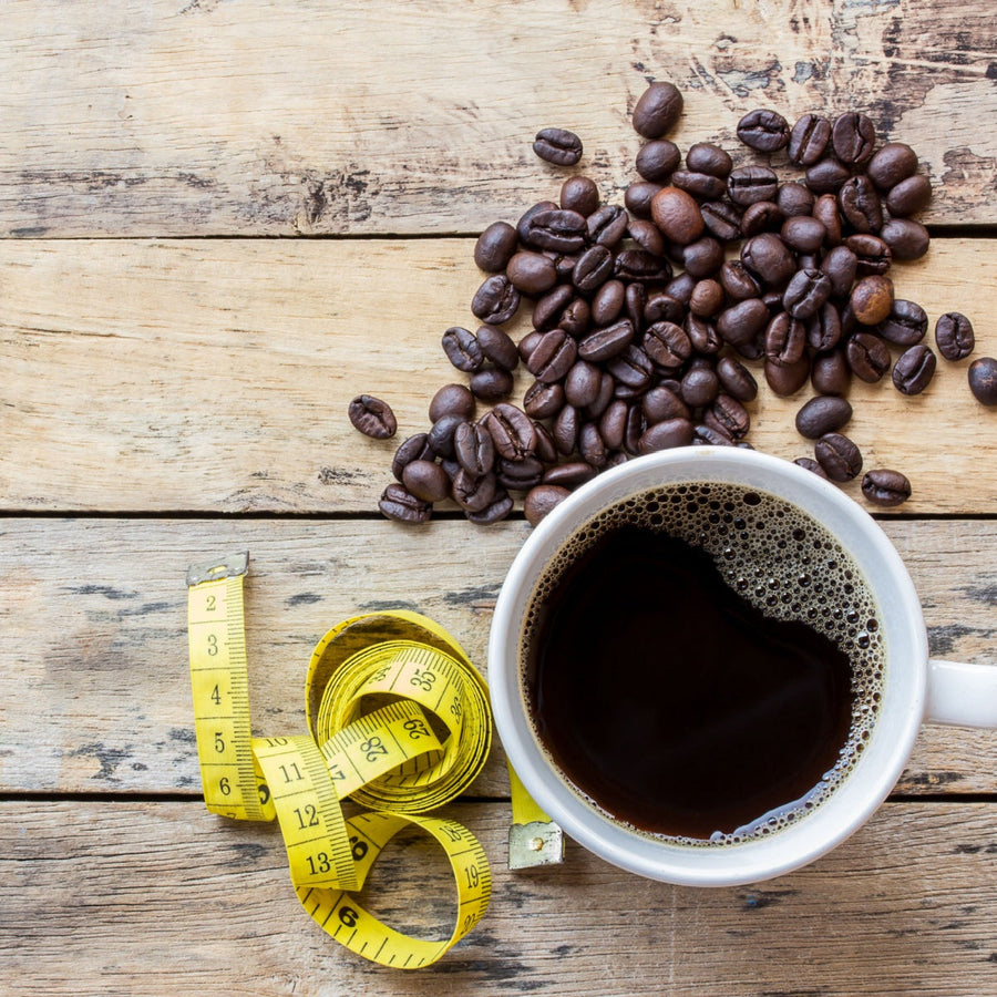 A white coffee mug filled with black coffee sits on a wooden table next to scattered coffee beans and a yellow measuring tape.