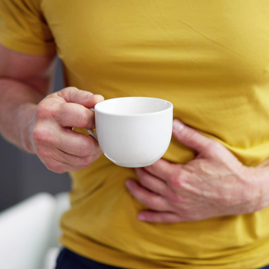 A person in a yellow shirt holds a white cup in one hand while gently pressing their other hand against their stomach, suggesting discomfort or a need for comfort in a casual indoor setting.