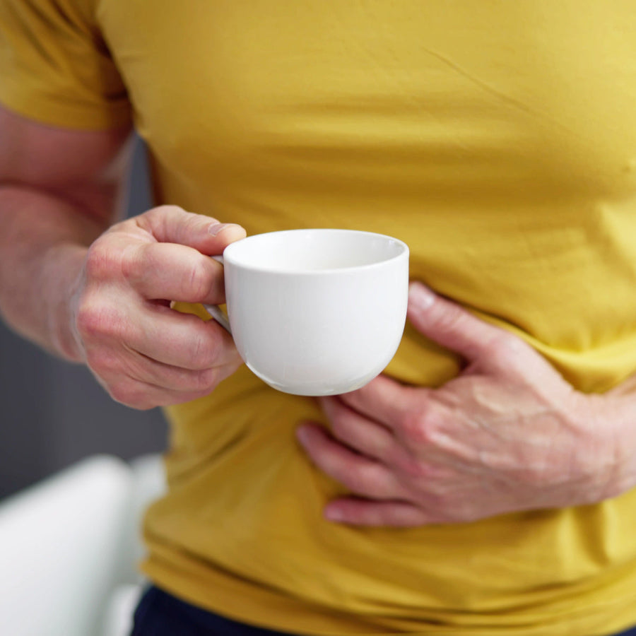 A person in a yellow shirt holds a white cup in one hand while gently pressing their other hand against their stomach, suggesting discomfort or a need for comfort in a casual indoor setting.