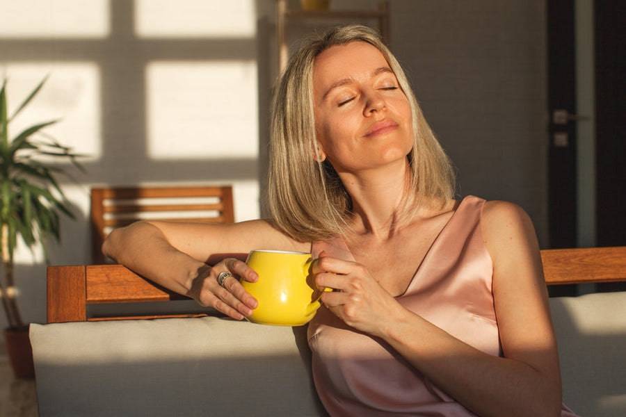 A woman with shoulder-length blonde hair sits on a couch, holding a yellow mug and smiling softly, enjoying sunlight streaming through a window in a cozy, modern living space.