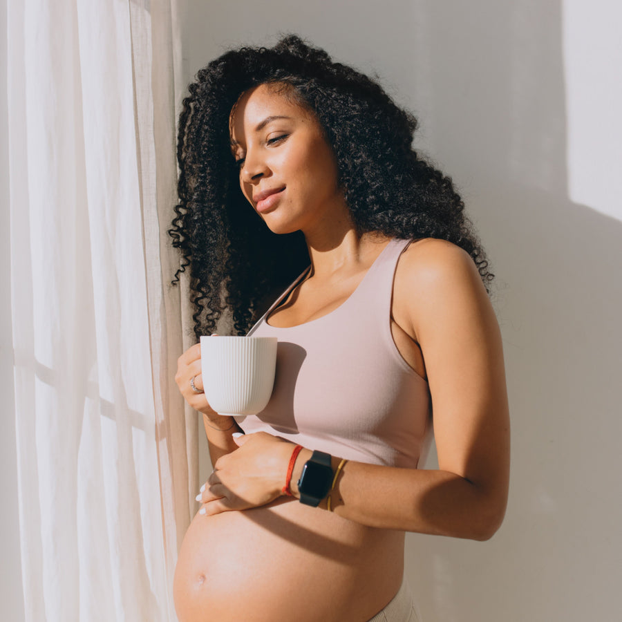 A pregnant woman with curly hair stands by a window, holding a white mug and gently cradling her belly, with a serene expression on her face as sunlight streams in.