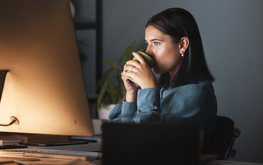 A woman sits at a desk in a dimly lit room, holding a green mug close to her face while gazing thoughtfully at a computer screen.