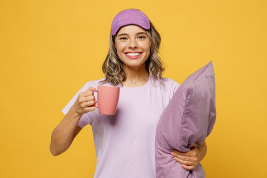 A smiling woman in a purple sleep mask and light purple shirt holds a pink mug in one hand and a lavender pillow in the other, set against a bright yellow background.