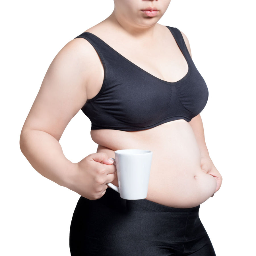 A person in a black sports bra and leggings stands holding a white mug, with a neutral expression and a visible belly, against a plain white background.