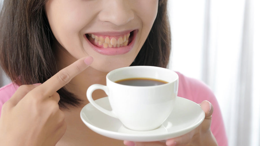 A woman with a bright smile points to her teeth while holding a white cup of coffee on a saucer, with soft natural light illuminating the background.