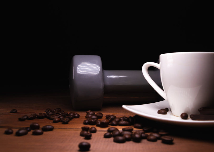 A gray dumbbell lies on a wooden surface next to a white coffee cup on a plate, with scattered coffee beans around, creating a contrast against a dark background.