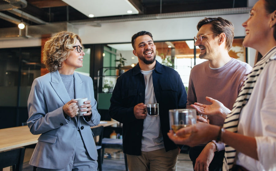 Four colleagues, smiling and engaged in conversation, stand in a modern office space, holding cups and glasses, enjoying a casual moment together.