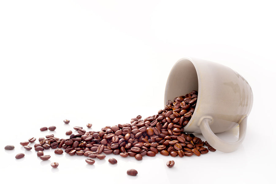 A gray coffee cup is tipped over, spilling a cascade of dark roasted coffee beans onto a white surface.