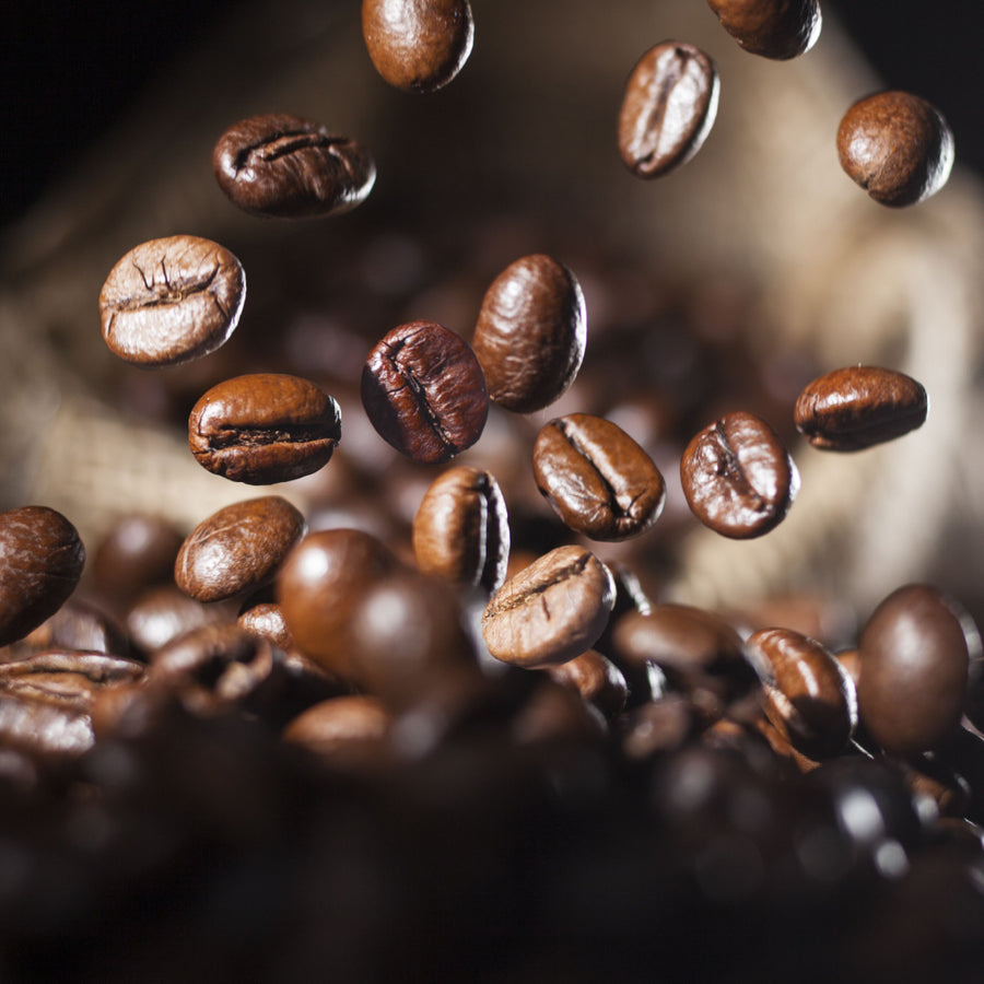Brown coffee beans are suspended mid-air above a pile of beans, with a textured burlap sack in the background, creating a dynamic scene of coffee preparation.