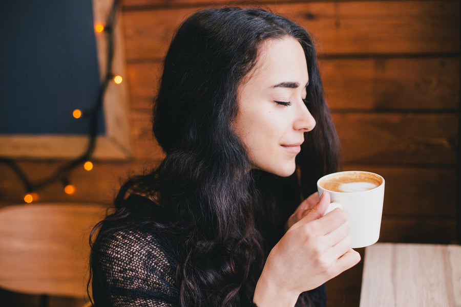 A woman with long dark hair, eyes closed, smiles gently while holding a cup of coffee in a cozy, wooden cafe setting with warm string lights in the background.