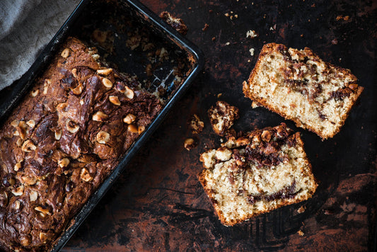 A freshly baked loaf of chocolate hazelnut bread sits in a dark baking dish, with two slices cut and placed beside it, revealing a marbled interior and scattered hazelnuts on top.