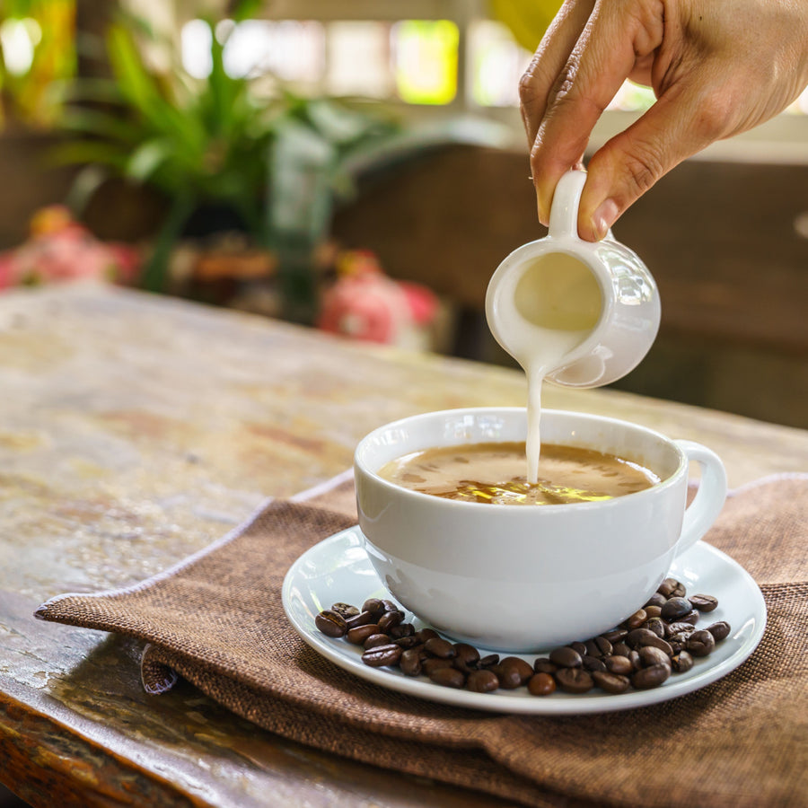 A hand pours cream from a small pitcher into a white coffee cup, resting on a saucer surrounded by coffee beans, with a blurred background of greenery and soft lighting.