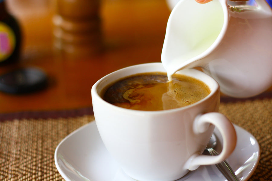 A hand pours cream from a small pitcher into a white coffee cup, creating swirls in the dark beverage, set on a textured placemat with a spoon beside it.
