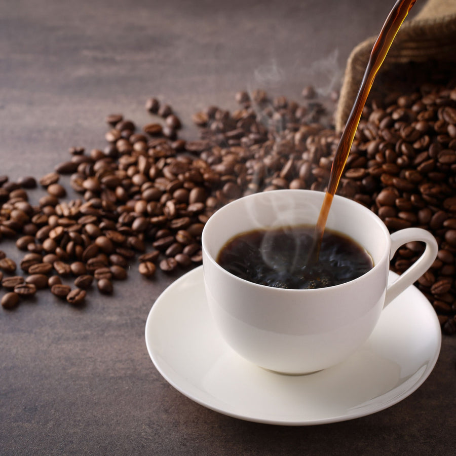 Hot coffee is being poured into a white cup on a saucer, surrounded by scattered coffee beans on a dark surface, with steam rising from the freshly brewed beverage.