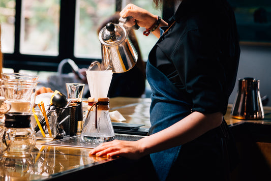 A barista in a black shirt and denim apron pours hot water from a kettle into a coffee filter set over a glass carafe, with various coffee-making tools visible on a polished countertop in a café.