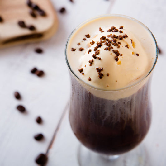 A tall glass filled with dark coffee topped with a scoop of vanilla ice cream and sprinkled with chocolate shavings, surrounded by scattered coffee beans on a light wooden surface.
