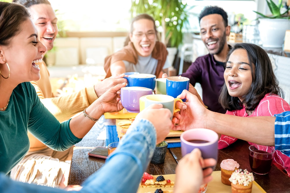 A group of six diverse friends joyfully toast colorful mugs while sitting around a table filled with pastries and snacks in a bright, lively café setting.