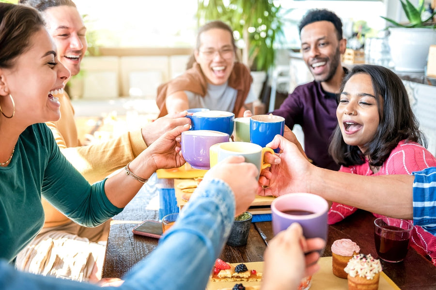 A group of six diverse friends joyfully toast colorful mugs while sitting around a table filled with pastries and snacks in a bright, lively café setting.