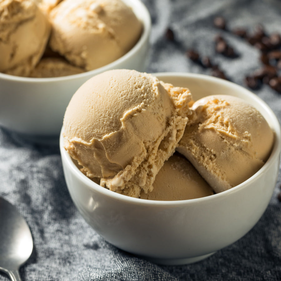Two bowls of creamy, light brown ice cream sit on a textured gray cloth, with a spoon nearby and scattered coffee beans in the background.