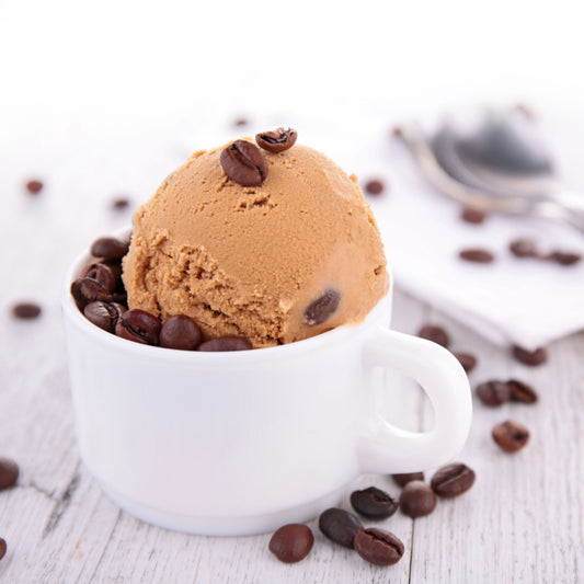 A scoop of coffee-flavored ice cream sits in a white cup surrounded by scattered coffee beans on a light wooden surface.