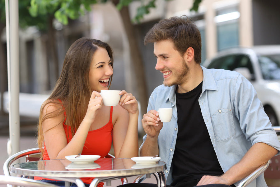 A young woman in a red tank top and a young man in a black shirt share a joyful moment over coffee at an outdoor café, smiling and laughing while holding their cups.