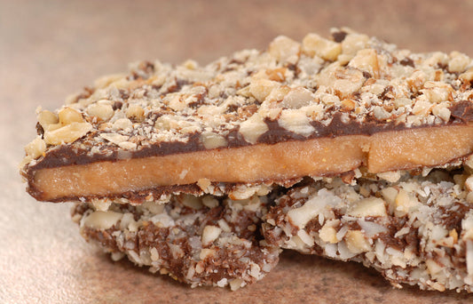 A close-up of a chocolate-covered candy bar with a layer of caramel, topped with crushed nuts, resting on a textured surface.