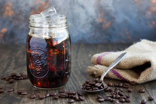 A glass jar filled with iced coffee and ice cubes sits on a wooden table, surrounded by scattered coffee beans and a silver spoon resting on a textured cloth.