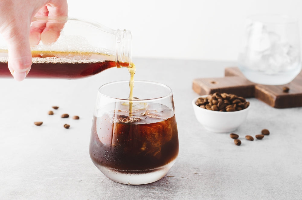 A hand pours cold brew coffee from a glass bottle into a round glass filled with ice, with coffee beans scattered on a light gray surface and a small bowl of beans nearby.