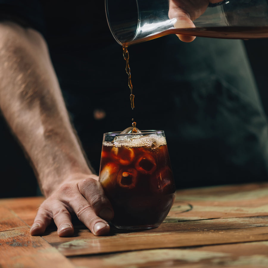 A hand pours dark liquid into a glass filled with ice, while another hand rests on the wooden table, creating a casual coffee-drinking scene against a dark background.