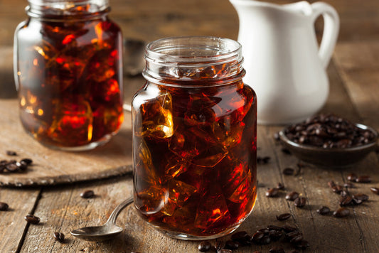 Two jars filled with dark coffee jelly sit on a wooden surface, accompanied by a small bowl of coffee beans and a white creamer pitcher in the background.