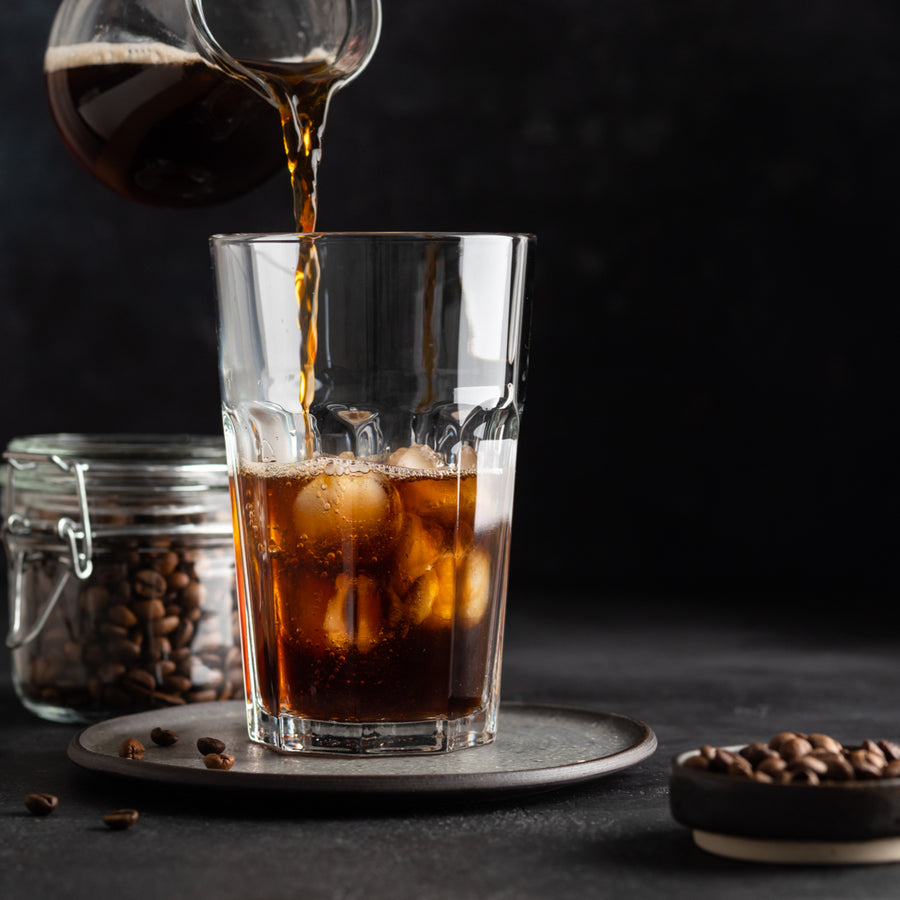 A glass filled with ice is being poured with dark coffee, while a jar of coffee beans and a small dish of whole coffee beans sit on a dark surface.