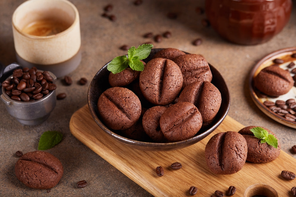 A bowl of chocolate cookies shaped like coffee beans is surrounded by coffee beans and a cup of espresso, with fresh mint leaves adding a pop of color on a wooden cutting board.