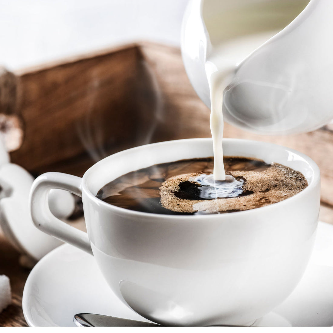 A stream of milk is being poured into a steaming cup of black coffee, placed on a saucer, with a rustic wooden background enhancing the cozy atmosphere.