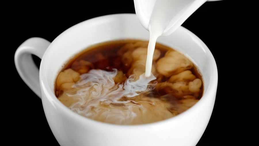 A stream of milk is being poured into a white cup of black coffee, creating swirling patterns in the liquid against a dark background.