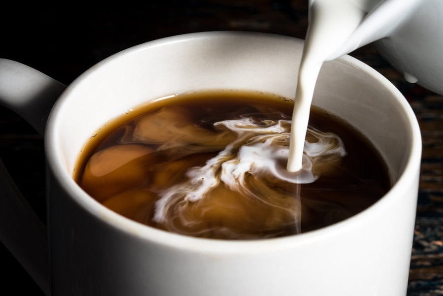 A stream of milk is being poured into a cup of black coffee, creating swirling patterns in the liquid against a dark wooden background.