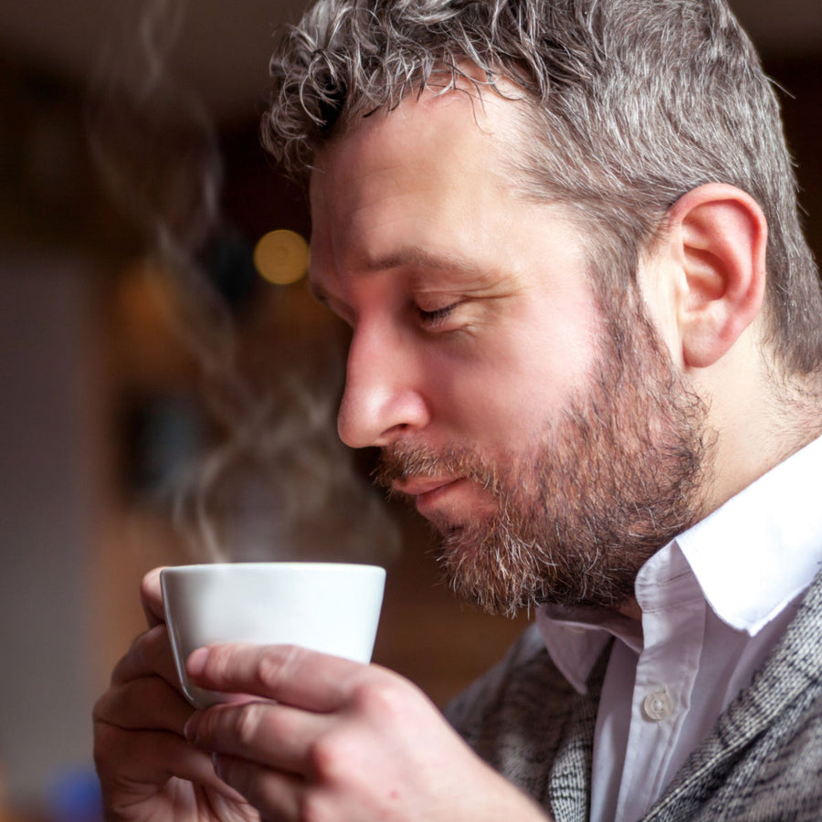 A man with short gray hair and a beard gently holds a steaming white cup, eyes closed and smiling slightly, enjoying a warm beverage in a cozy, softly lit setting.
