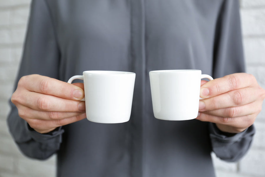 A person in a gray shirt holds two small white cups, one in each hand, against a light brick background, suggesting a moment of sharing or offering.