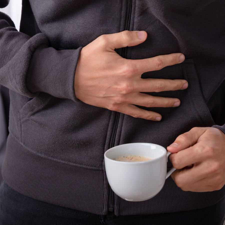 A person in a dark hoodie holds a white cup of coffee in one hand while placing the other hand on their stomach, suggesting discomfort or a casual moment.