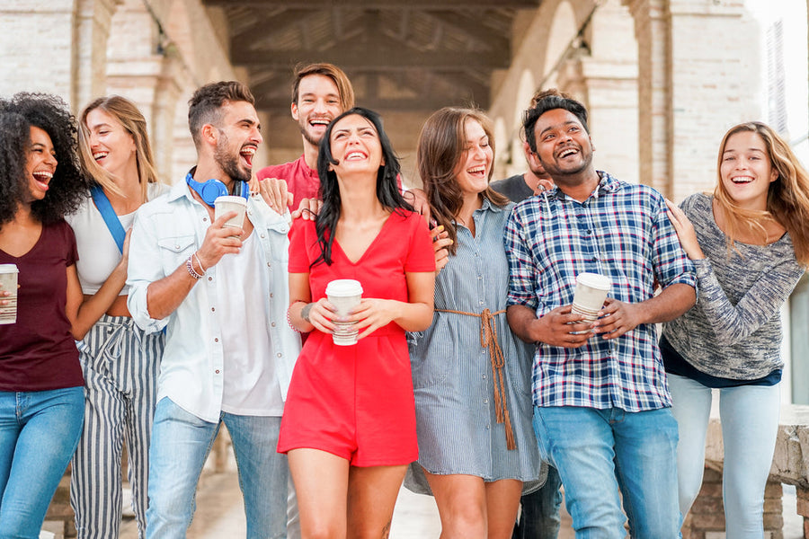 A diverse group of eight young adults joyfully laugh and walk together, holding coffee cups, under an arched walkway in a sunny outdoor setting.