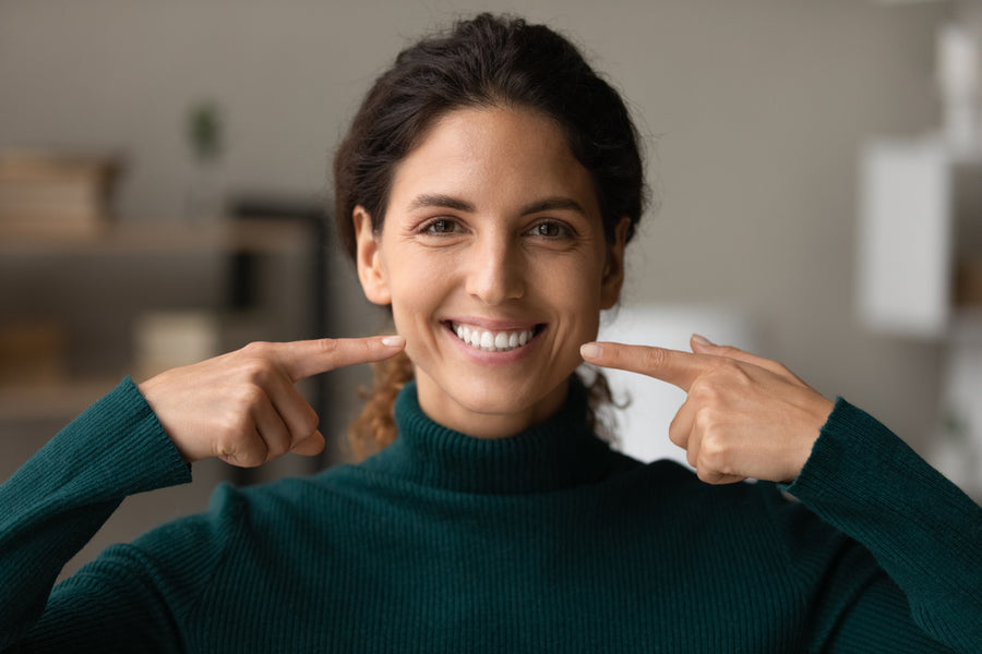 A woman in a green sweater smiles brightly while pointing to her own cheeks, set against a softly blurred indoor background.