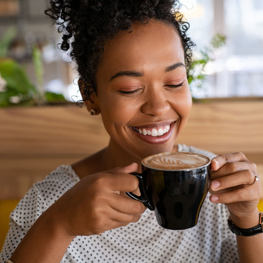 A woman with curly hair smiles joyfully while holding a black coffee cup, enjoying a moment in a bright café filled with greenery.