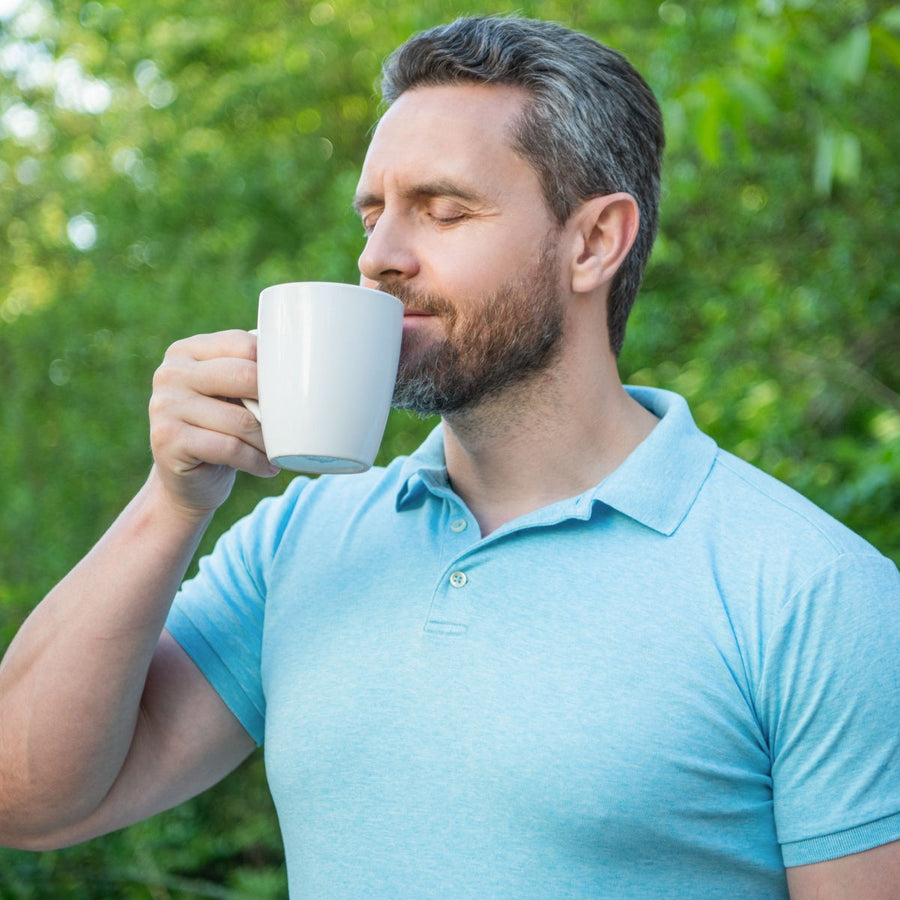 A man with a beard in a light blue polo shirt enjoys a warm drink, eyes closed and smiling, while standing outdoors surrounded by greenery.